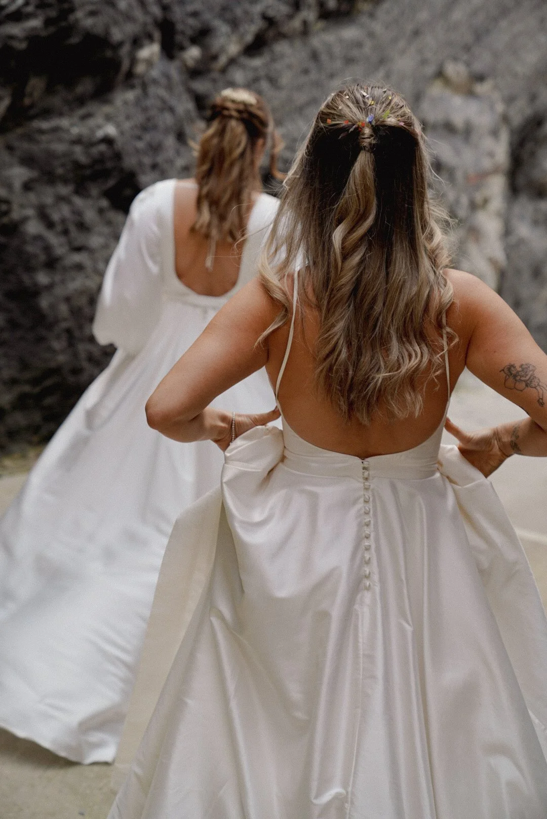 Two women in wedding dresses standing on a sandy surface near dark rocky cliffs, one woman with her back to the camera and the other facing away, adjusting her dress.