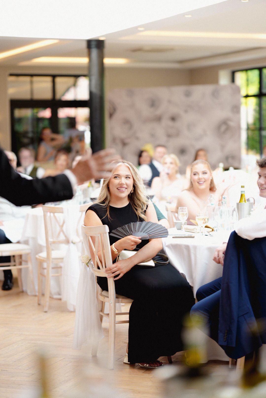 People at a formal event, seated at tables with white tablecloths, in a well-lit venue with large windows, listening to a speaker.
