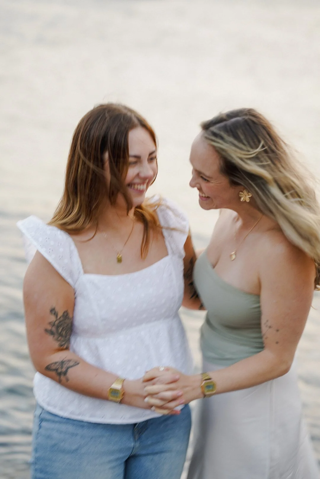 Two women smiling and holding hands near a body of water during sunset.