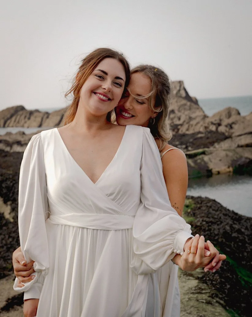 Two women, one in a white dress and the other in a sleeveless top, smiling and embracing on a rocky coastal background.