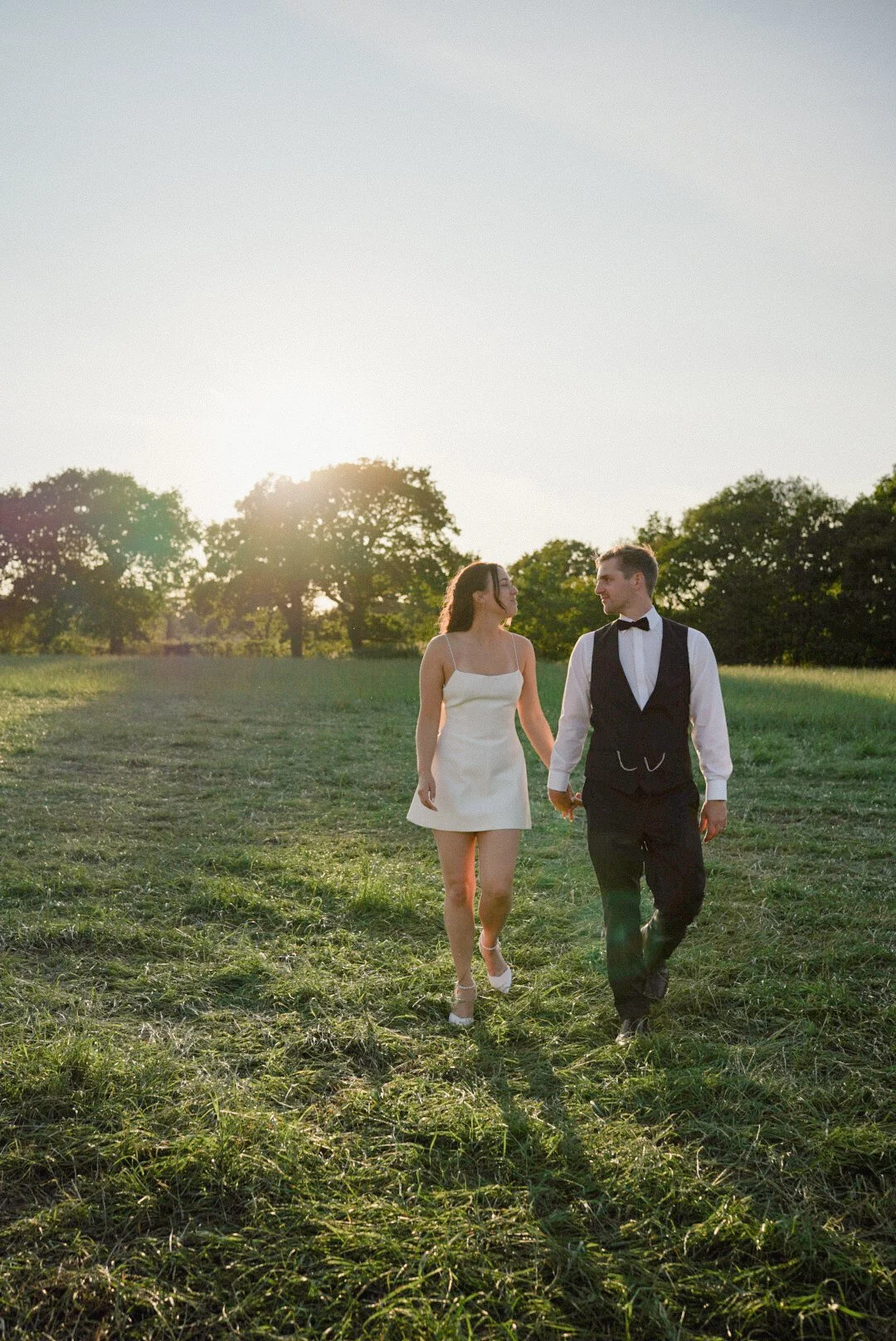 A bride and groom walking hand in hand in a grassy field with trees and the setting sun in the background.