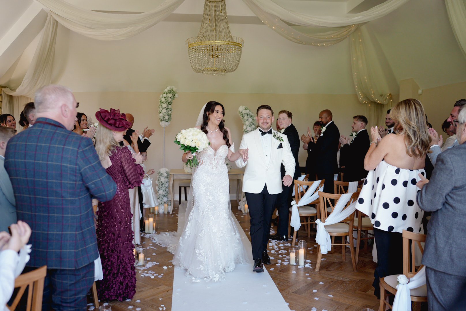 Bride and groom walking down the aisle after wedding ceremony, surrounded by family and friends applauding inside a decorated wedding venue.