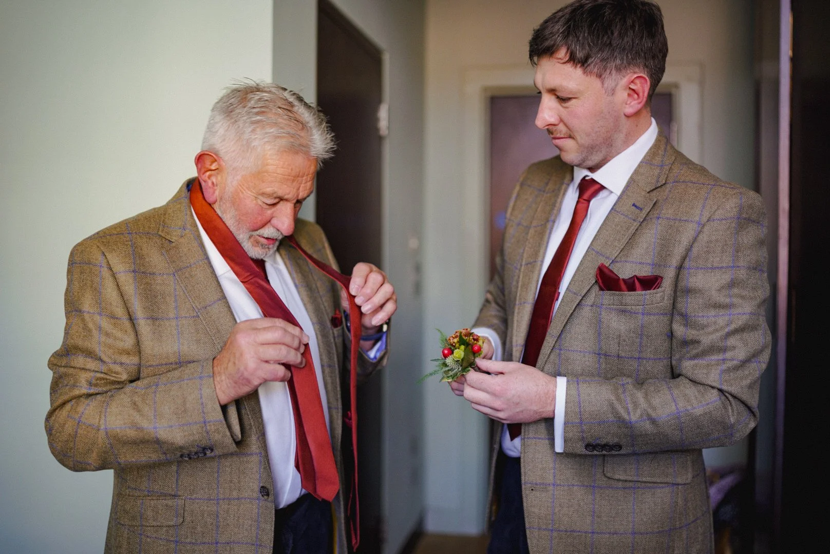 Two men in brown plaid suits preparing for a formal event, with one holding a boutonniere and the other adjusting his red tie.