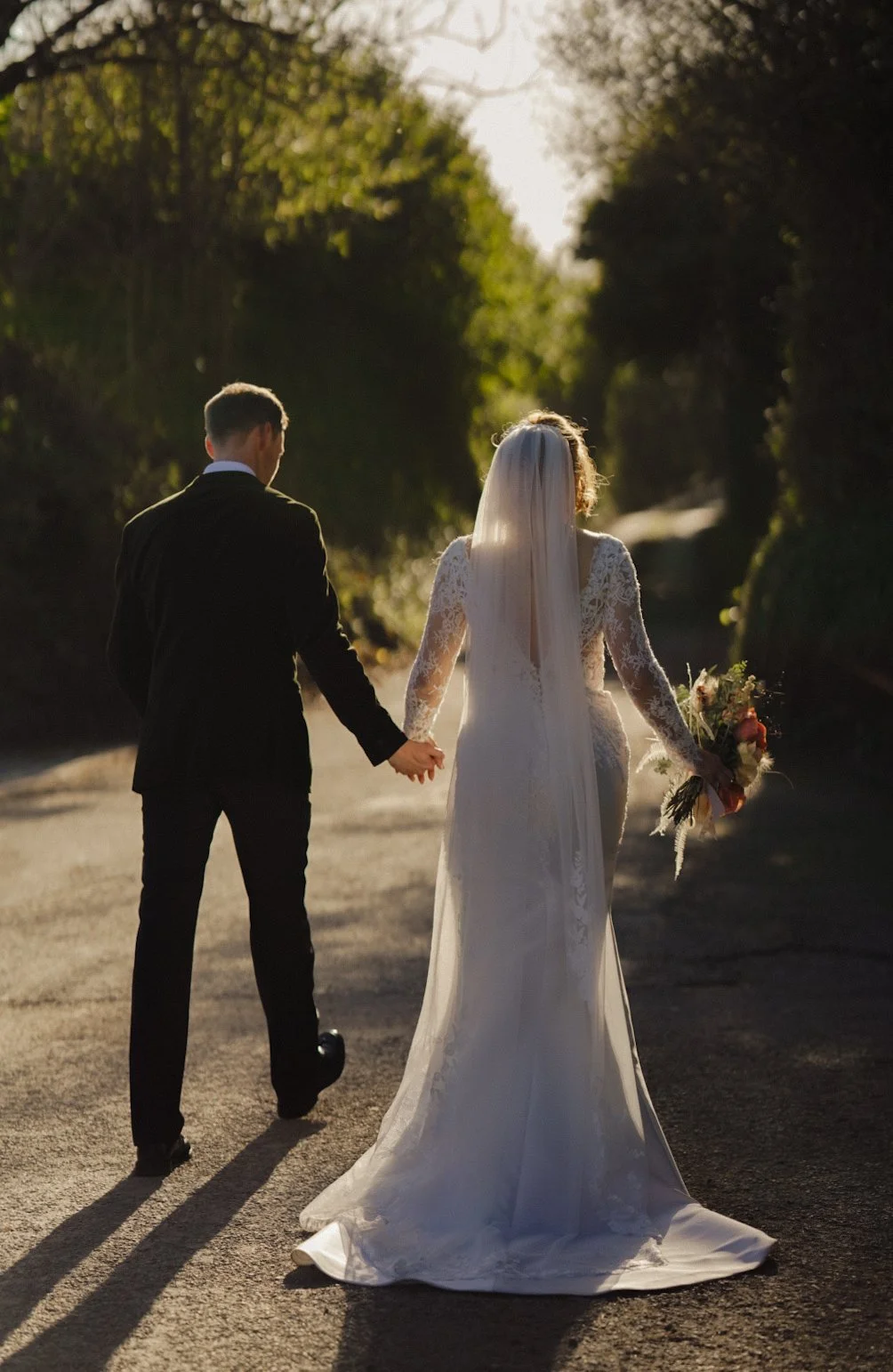 A bride and groom holding hands and walking on a tree-lined dirt road at sunset, with the bride wearing a white lace wedding dress and veil, and the groom in a black suit.