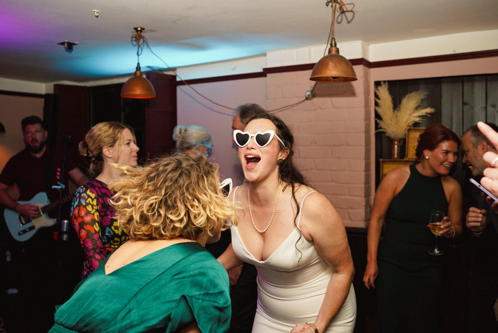 A group of people dancing and socializing at a party or celebration, with one woman wearing white dress and heart-shaped sunglasses singing or shouting joyfully.