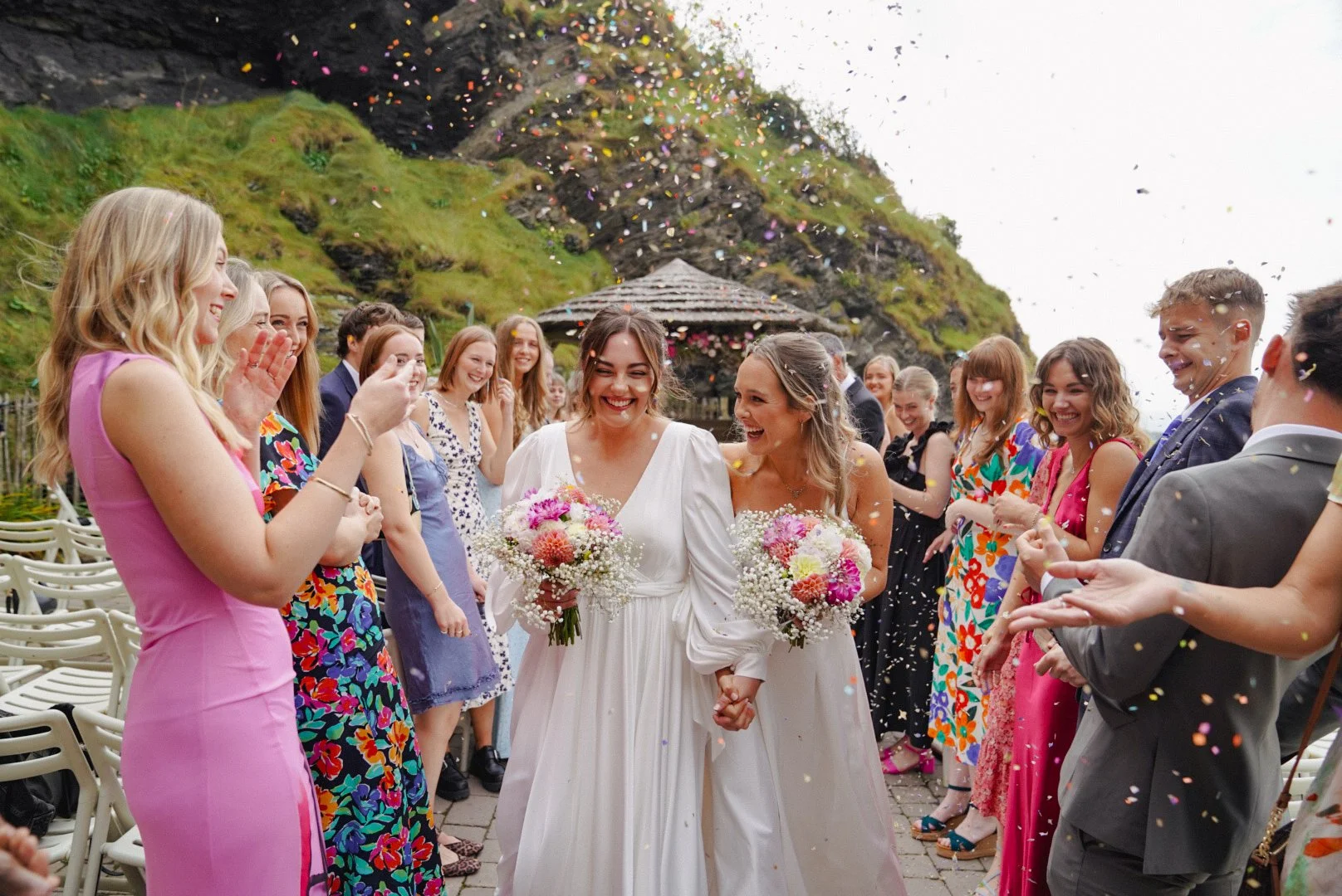 Two women in white wedding dresses holding hands and bouquet, walking through a celebratory gathering of friends and family outdoors, with confetti falling, green hillside, and wooden gazebo in background.