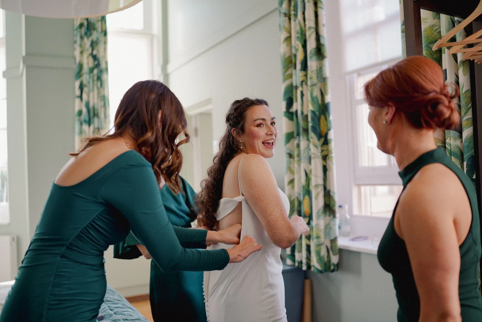 Bridesmaids helping bride into her wedding dress in a brightly lit room with green and yellow floral curtains.