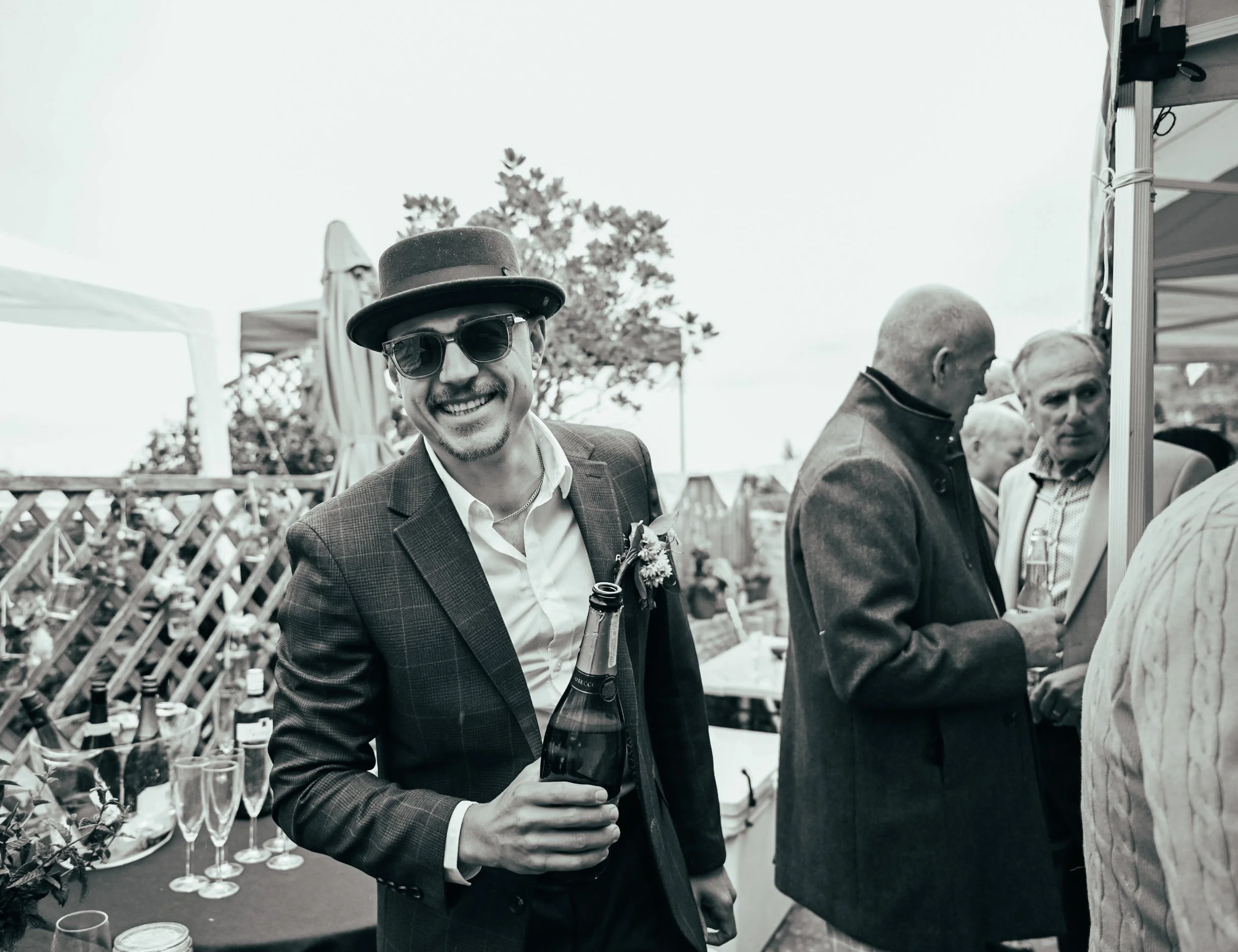 Man wearing sunglasses, a hat, and a suit holding a champagne bottle at an outdoor party