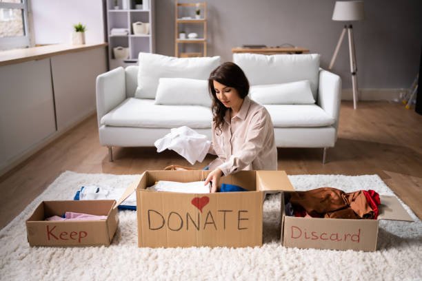 A woman sorting laundry in a living room with labeled boxes that read "Keep," "Donate," and "Discard."