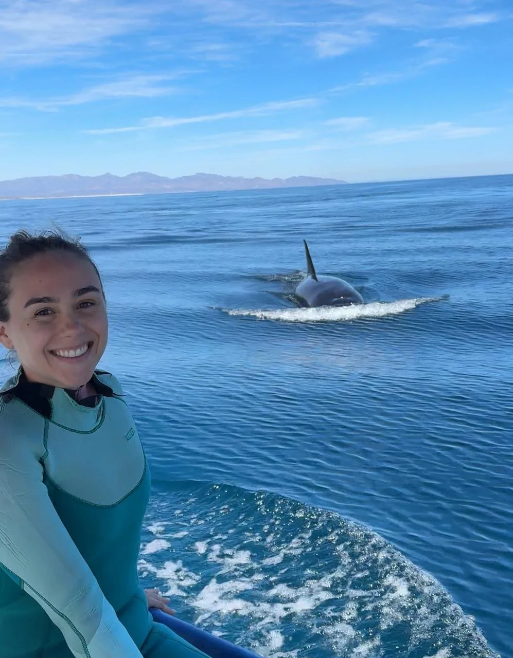 Mujer hermosa sonriendo en un bote en el mar con una orca a lo lejos, en un día soleado con cielo azul y algunas nubes.