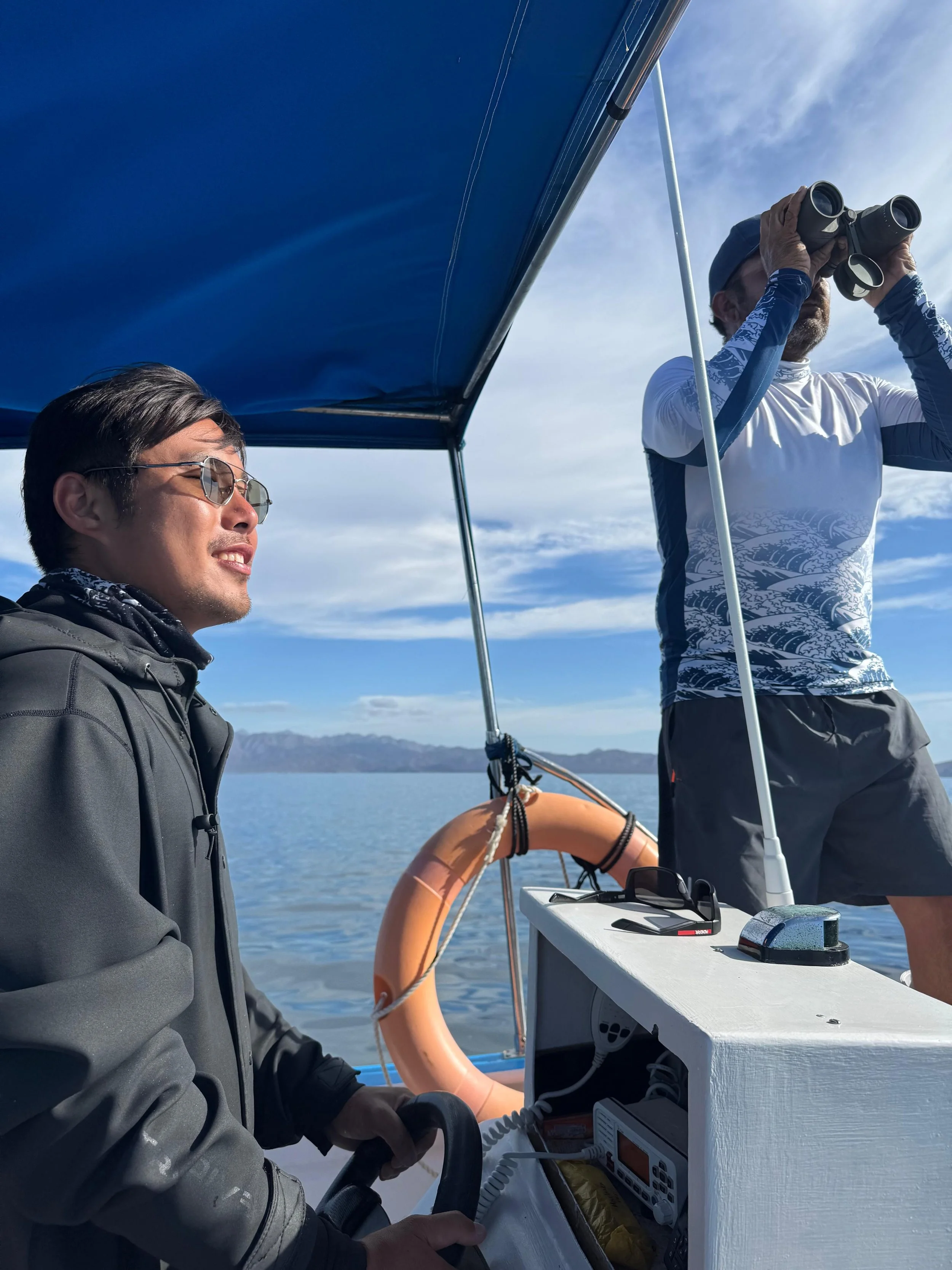 Dos hombres en un barco, uno con gafas de sol y el otro capitan con binoculares, observando el horizonte en un día despejado