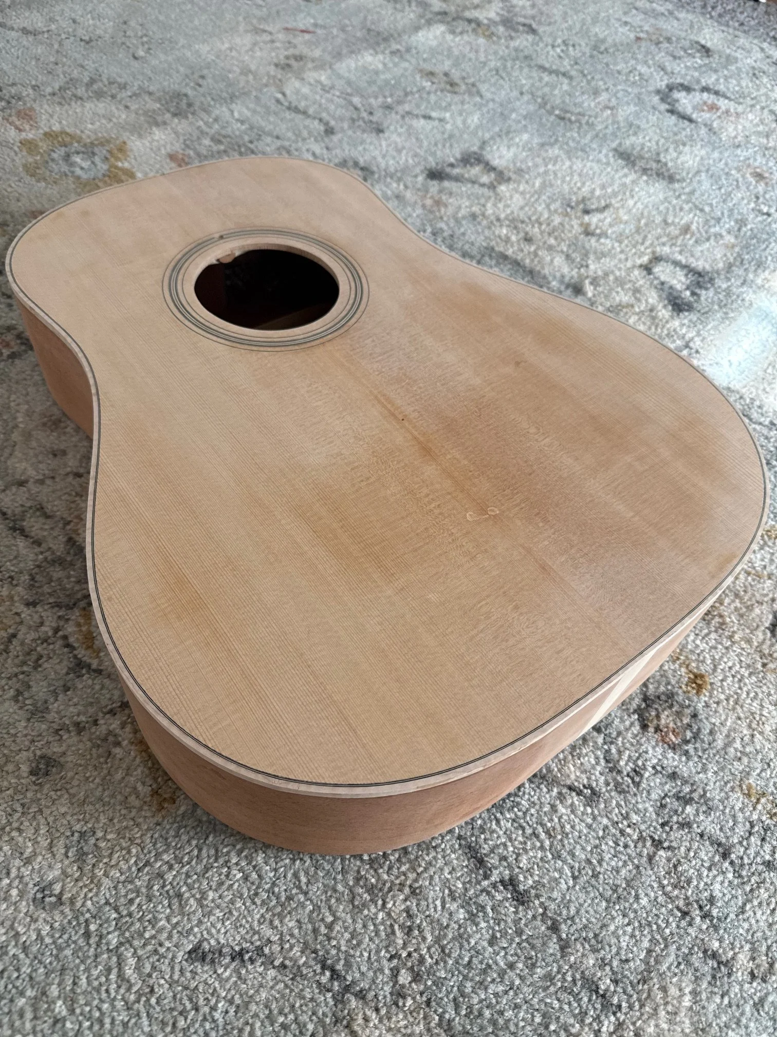 An unfinished wooden acoustic guitar lying on a textured carpet.