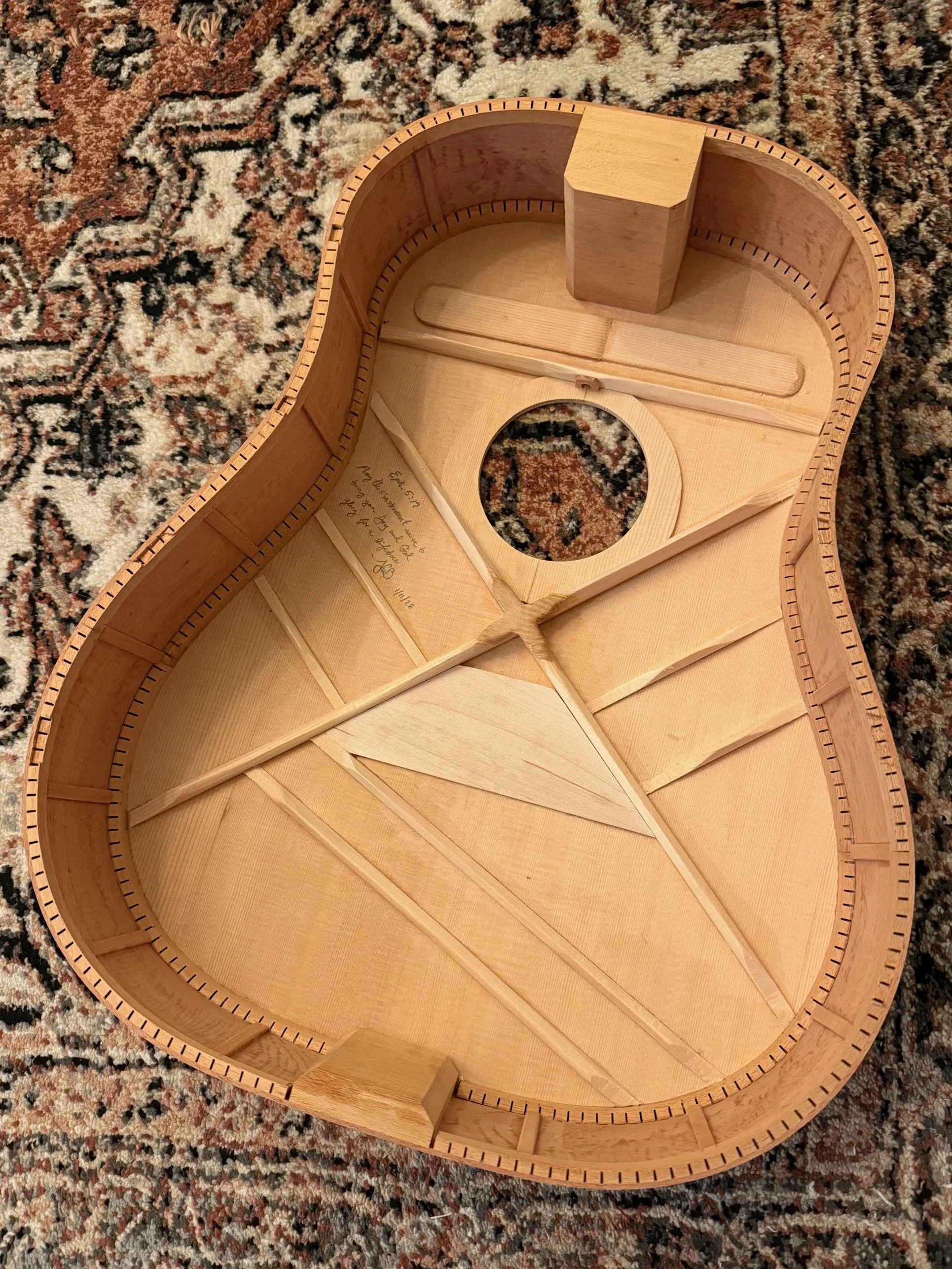 Inside of an unfinished wooden ukulele body with supporting braces and a circular sound hole, resting on a patterned rug.