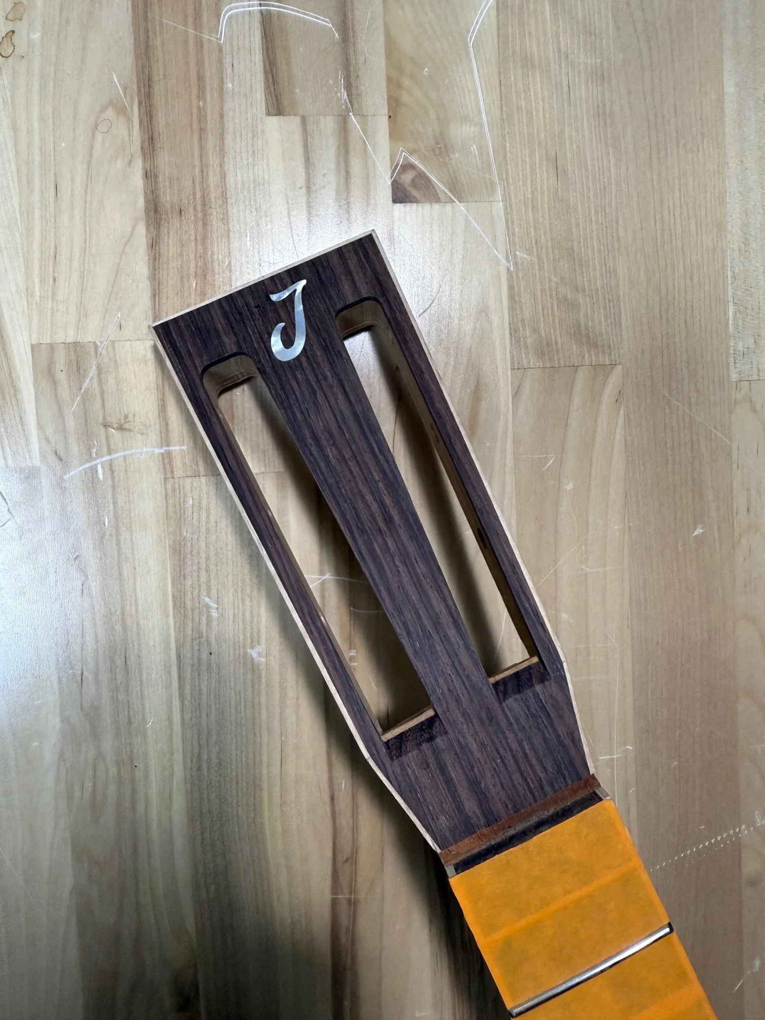 Wooden guitar headstock with a white letter J and a neck with orange tape at the top.