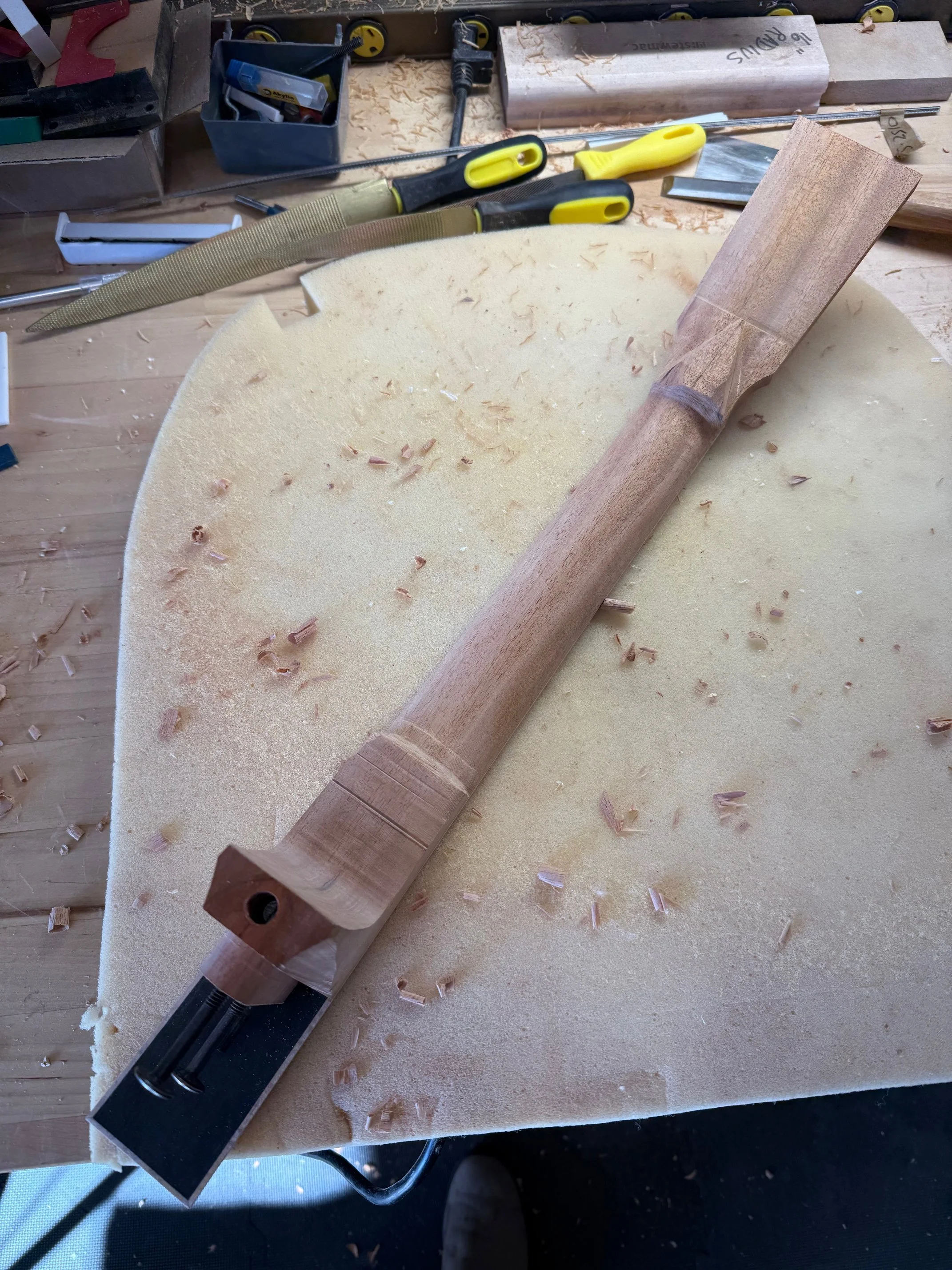 Unfinished wooden guitar neck on a workbench with woodworking tools and foam padding, surrounded by wood shavings.