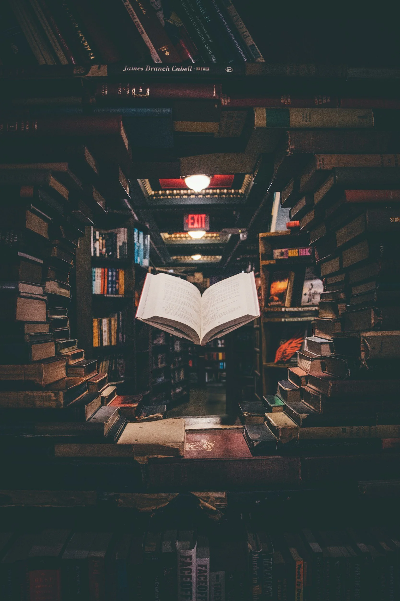 A floating open book in a dimly lit bookstore or library, surrounded by shelves filled with stacked books, with an exit sign glowing in the background.
