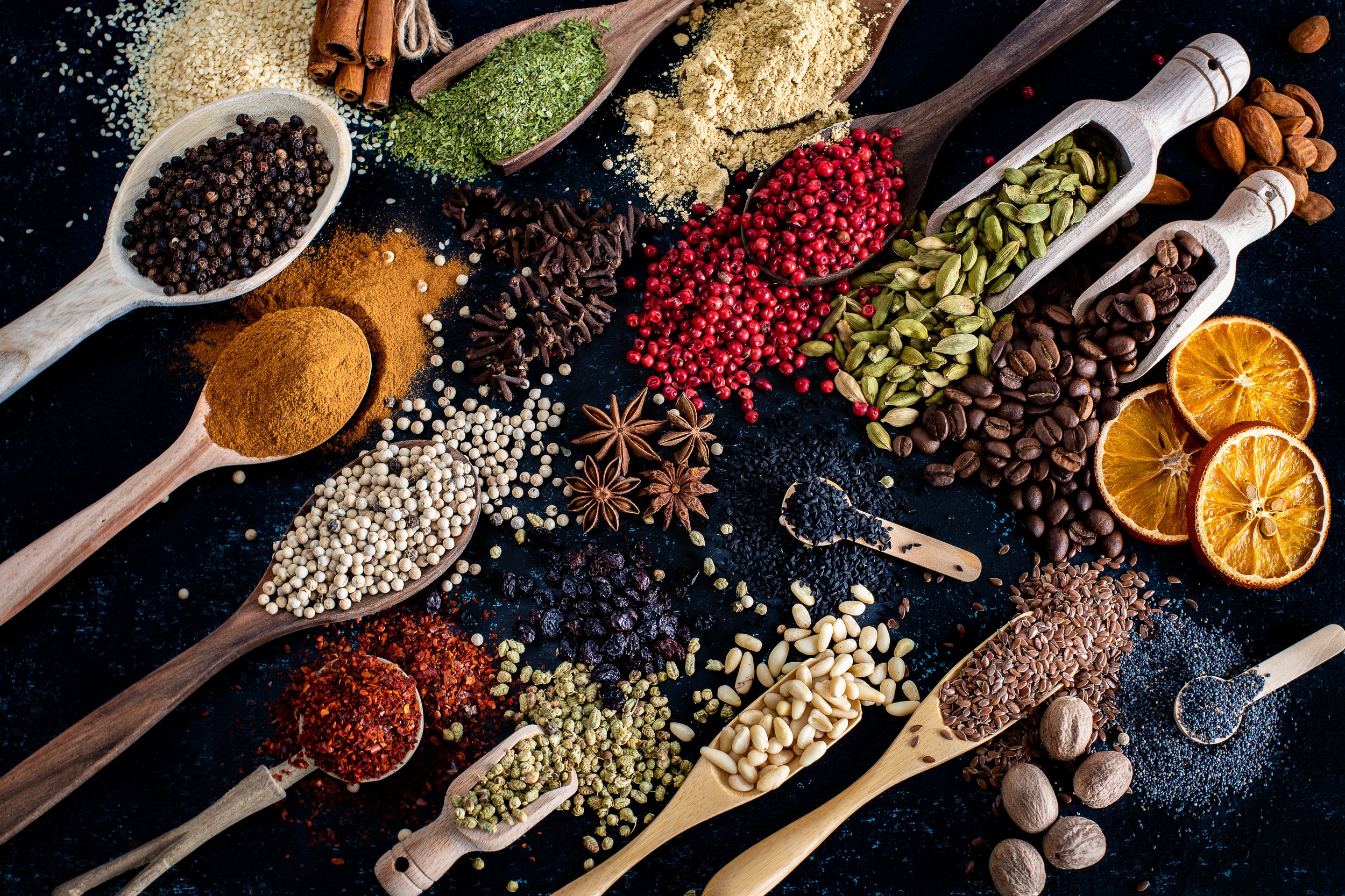 Assorted spices and herbs in spoons and scoops, including peppercorns, cinnamon sticks, dried herbs, star anise, and orange slices, on a dark background.