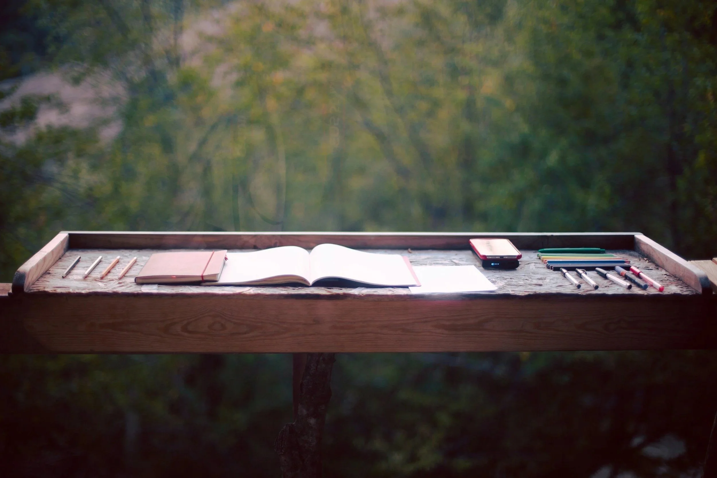 Bureau en bois avec un carnet, un livre ouvert, un téléphone portable, des stylos et des notes, sur une table en plein air avec un fond de forêt.