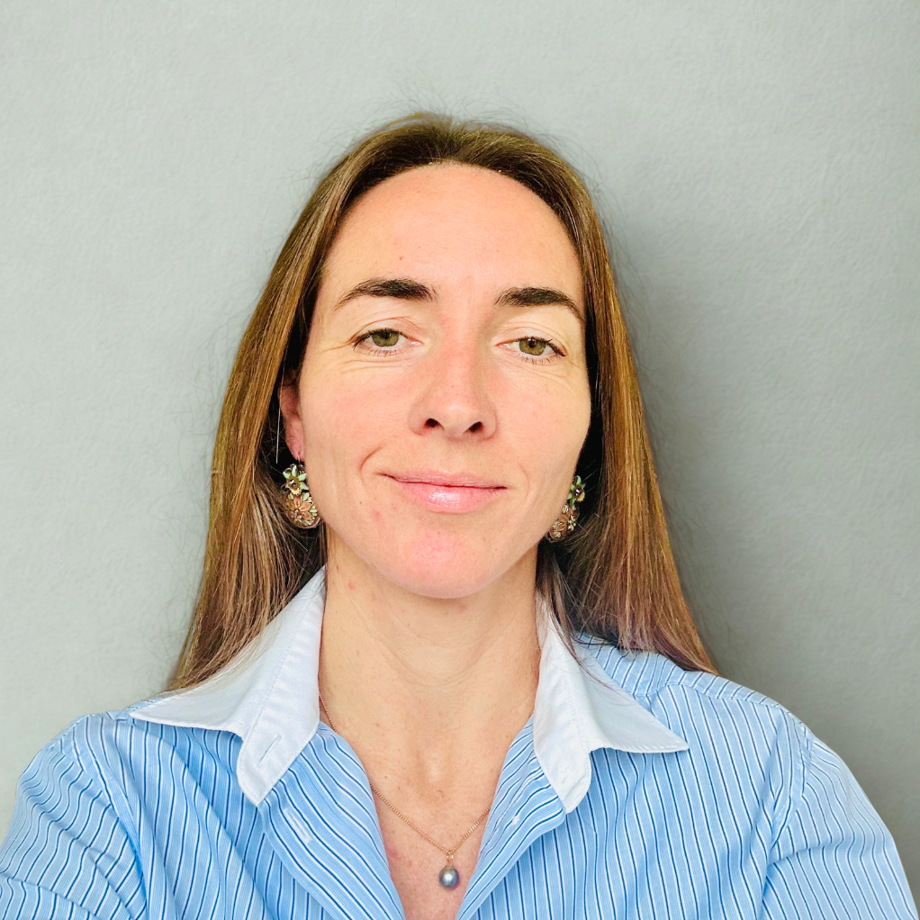 Une femme avec des cheveux bruns longs, portant une chemise à rayures bleues et blanches, avec des boucles d'oreilles et un collier, posant devant un mur gris.