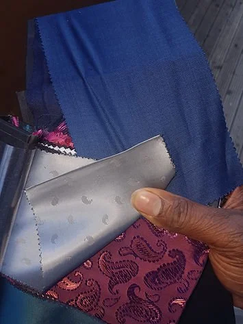A person holding a metallic cloth with water droplets, with fabric samples including blue, pink, and patterned purple in the background.