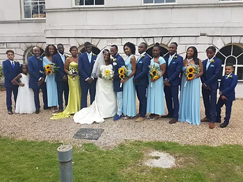 A wedding party outdoors with the bride and groom in the center, surrounded by bridesmaids and groomsmen, all dressed in blue and yellow. The group is posing in front of a white building with arched windows.