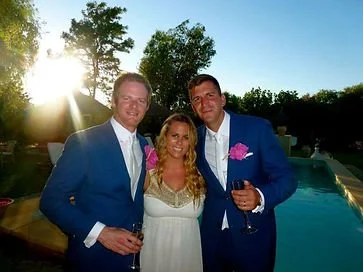 Three people, two men and a woman, dressed in formal attire, smiling outdoors near a swimming pool during sunset, holding wine glasses.