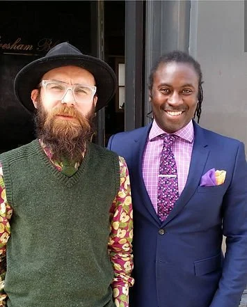 Two men standing outdoors, one wearing a black hat, glasses, a green vest, and a patterned shirt, the other in a blue suit with a purple tie and pocket square, both smiling.