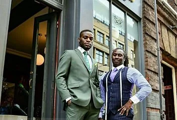 Two men standing outside a building, one in a suit and tie, the other in a vest and dress shirt, near a glass door.