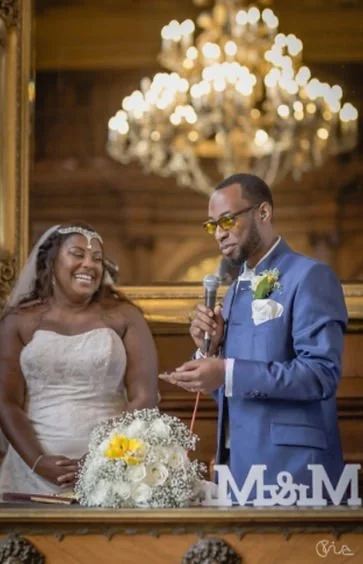 A bride and groom at a wedding ceremony, with the groom reading vows from a paper, in a church with a chandelier overhead.