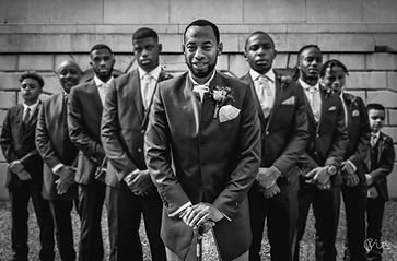 A group of men dressed in suits, with one man in front smiling, standing outdoors in front of a brick wall.