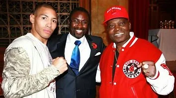 Three people standing together, smiling for the camera. The woman on the left is wearing a light-colored hoodie, the man in the middle is dressed in a suit with a blue tie, and the man on the right is wearing a red jacket and red cap with a logo.