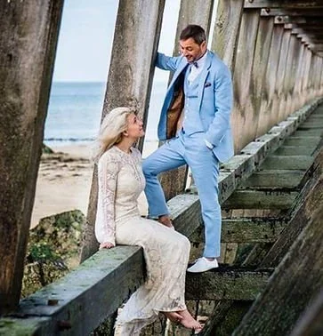 A man and woman on a beach, with the man standing on a wooden platform and the woman sitting on a beam, sharing a moment.