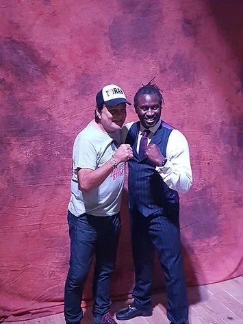 Two men standing together, one wearing a baseball cap and gray t-shirt, the other in a white shirt with a dark vest and tie, smiling and making a fist bump pose against a pinkish-brown textured background.