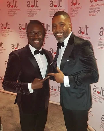 Two men in tuxedos at an event, smiling and pointing at each other, with a backdrop displaying the logo 'a-IT' and related text.