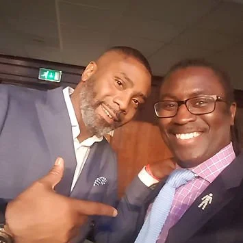Two men smiling, one pointing at the other, in a room with a wooden wall and an exit sign.