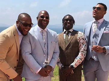 Four men dressed in suits standing outdoors near a body of water, smiling and laughing.