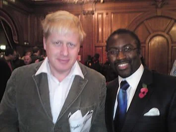 Two men in formal attire posing together at an indoor event, with wood-paneled walls in the background.