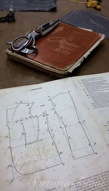 A sewing pattern book and a technical drawing of a garment piece on a table along with a pair of scissors, keys, and fabric swatches.