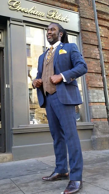 A man in a blue plaid suit, brown vest, and yellow tie standing outside a shop called Gresham Blake.