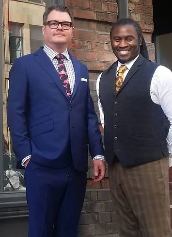 Two men in suits standing outside in front of a brick building, smiling at the camera.