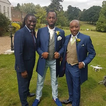 Three men dressed in suits standing outdoors on a grassy area, with trees and buildings in the background.