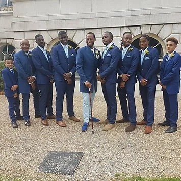 Group of nine men and boys dressed in blue suits posing outdoors in front of a stone wall.
