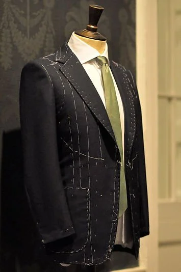 A mannequin dressed in a tailored dark suit with white pinstripes, white shirt, and a green tie, displayed in a store or showroom.