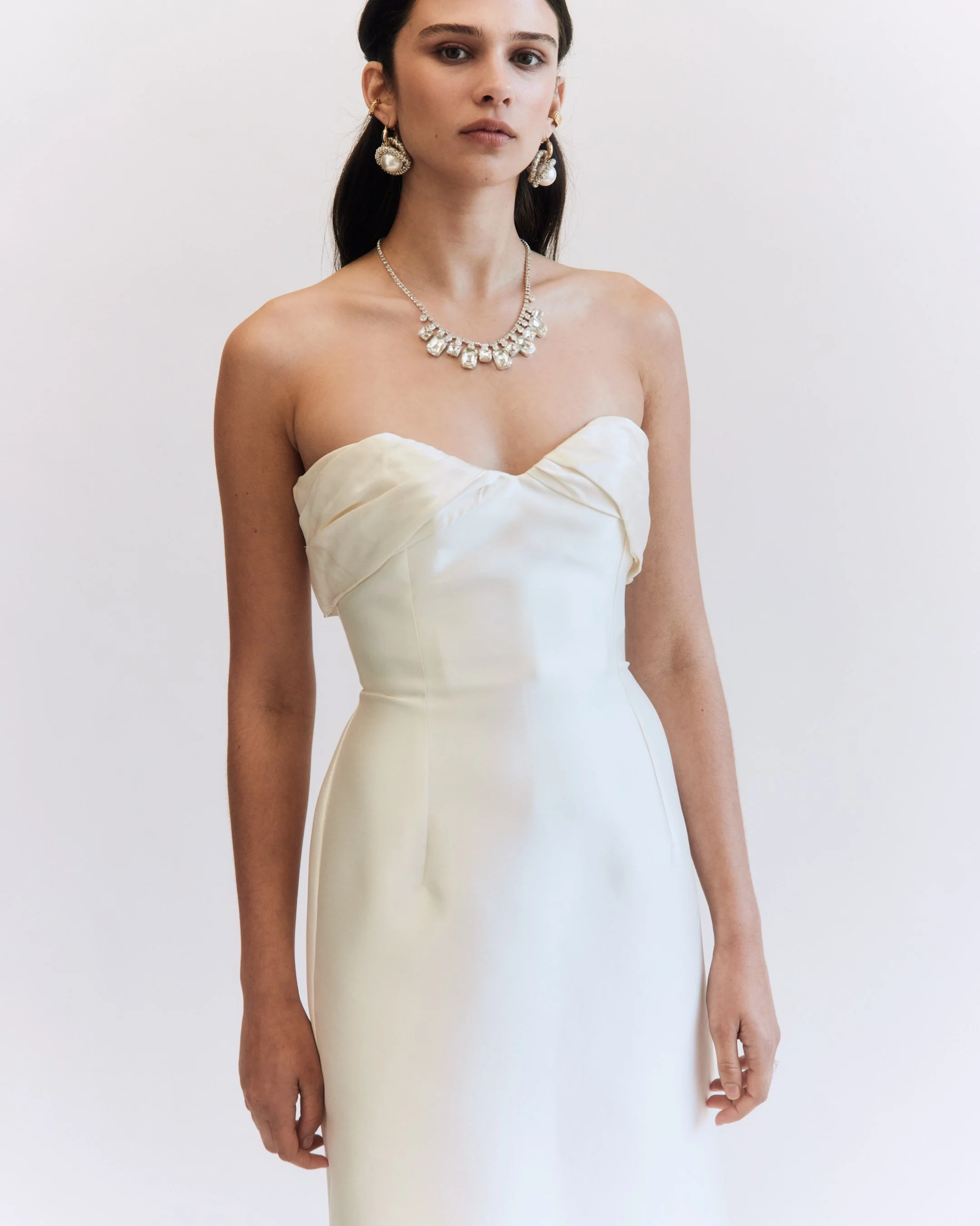 A woman in a strapless white wedding dress, wearing pearl and diamond jewelry, standing against a plain white background.