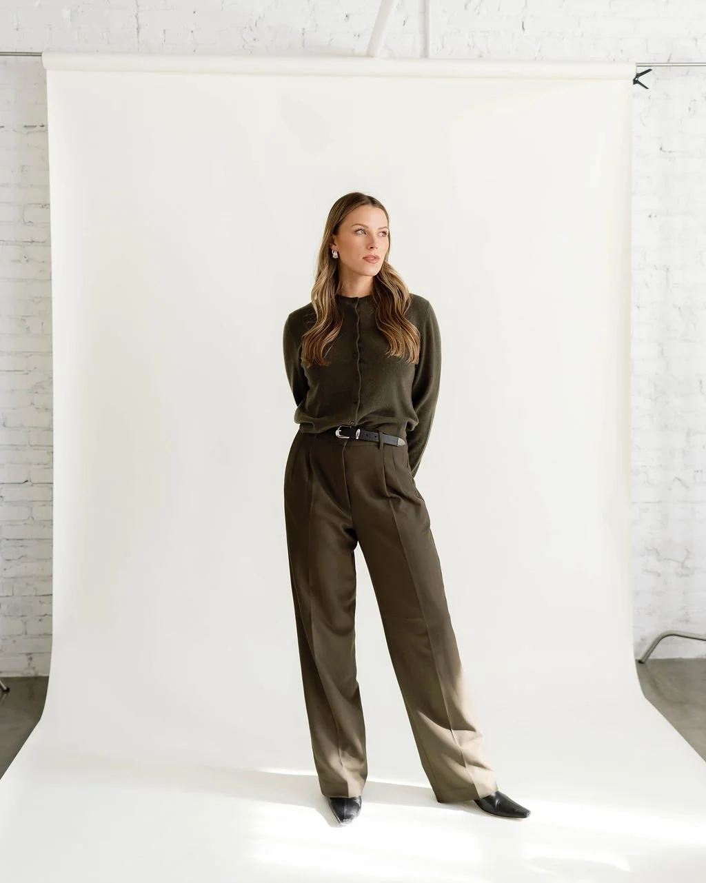 Woman in dark green blouse and brown pants standing against white backdrop in photo studio.