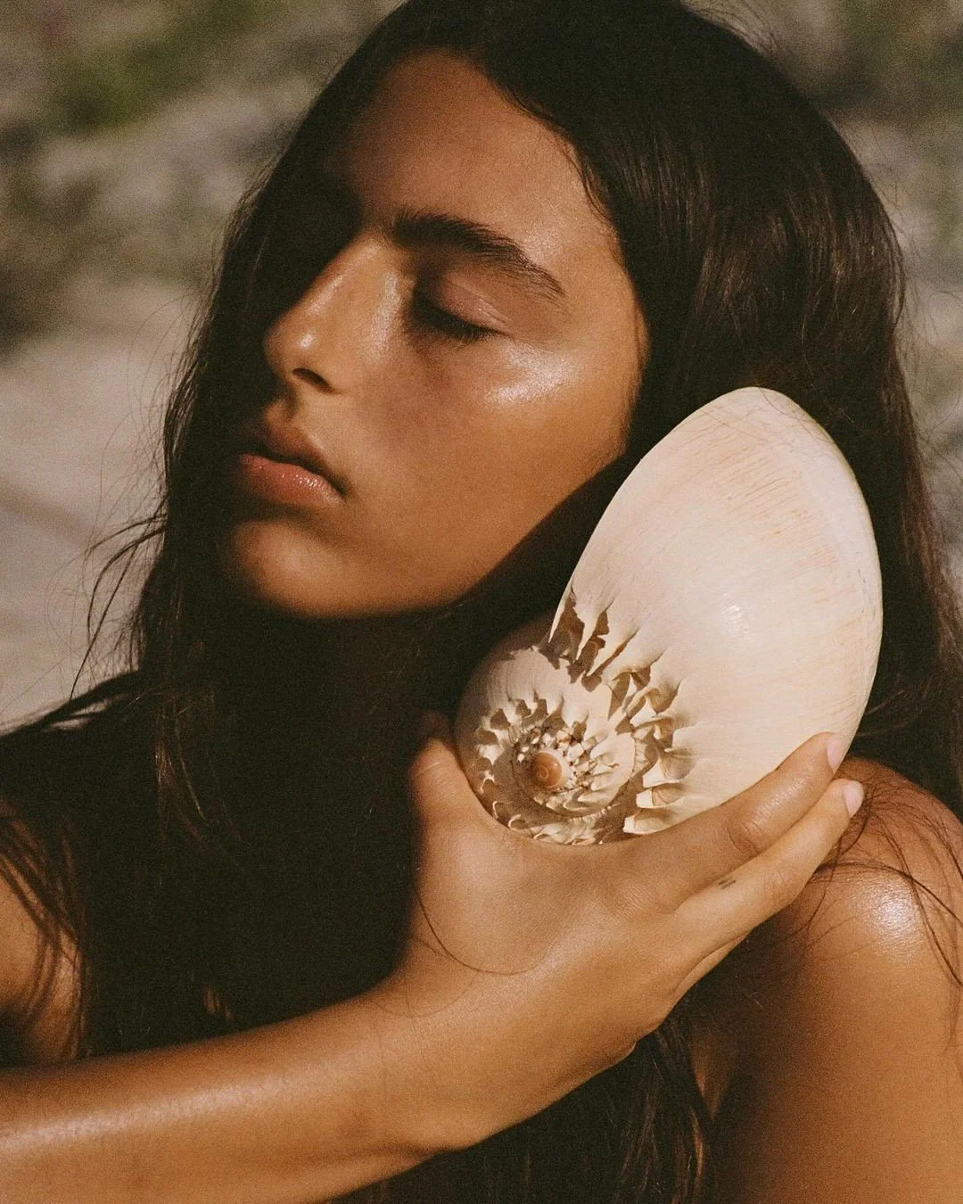 A woman with closed eyes holding a large seashell against her ear.