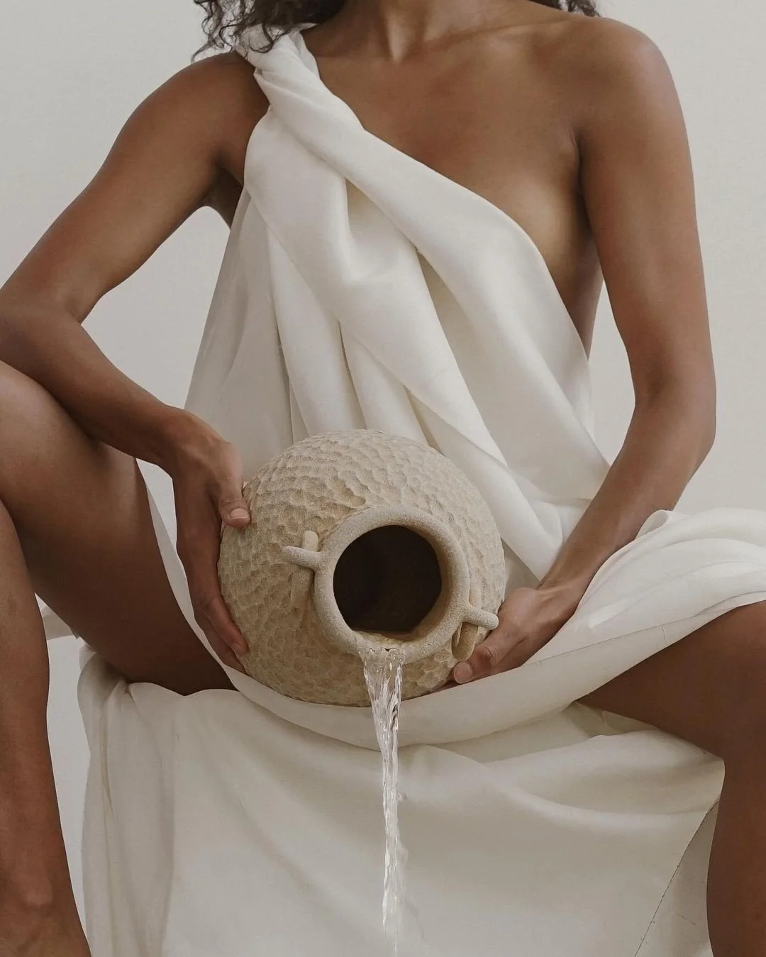 A person dressed in white fabric draped over shoulders and lap, holding a textured, round ceramic pitcher with a small handle, pouring water into their lap.