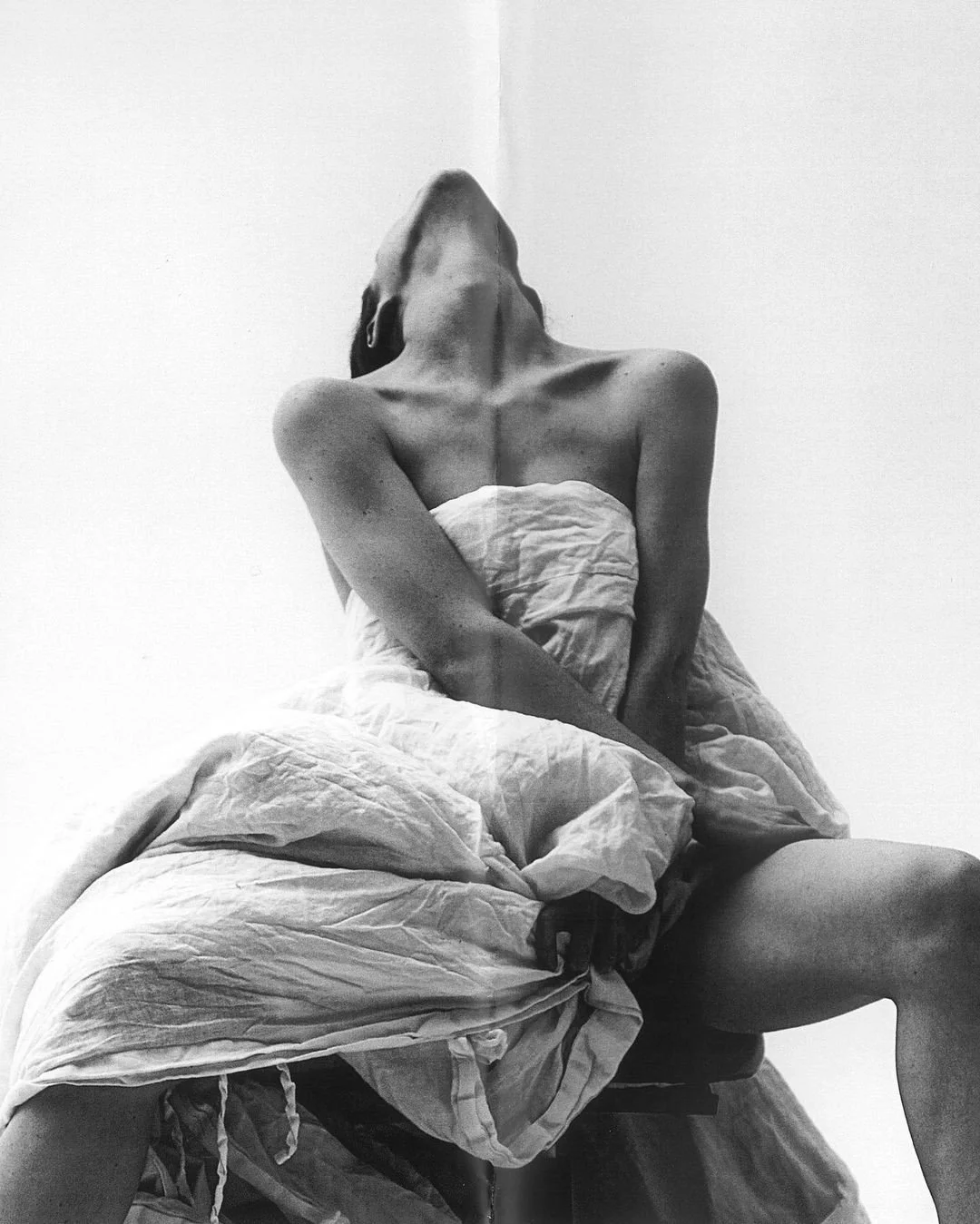 Black and white photo of a person sitting on a bed, wrapped in a rumpled sheet, with their head tilted back and arms crossed over their chest.