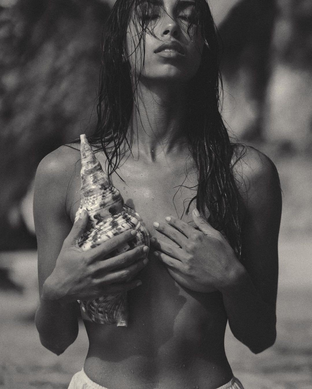 A woman with wet hair holding a large seashell and covering her chest with her hands on a beach.