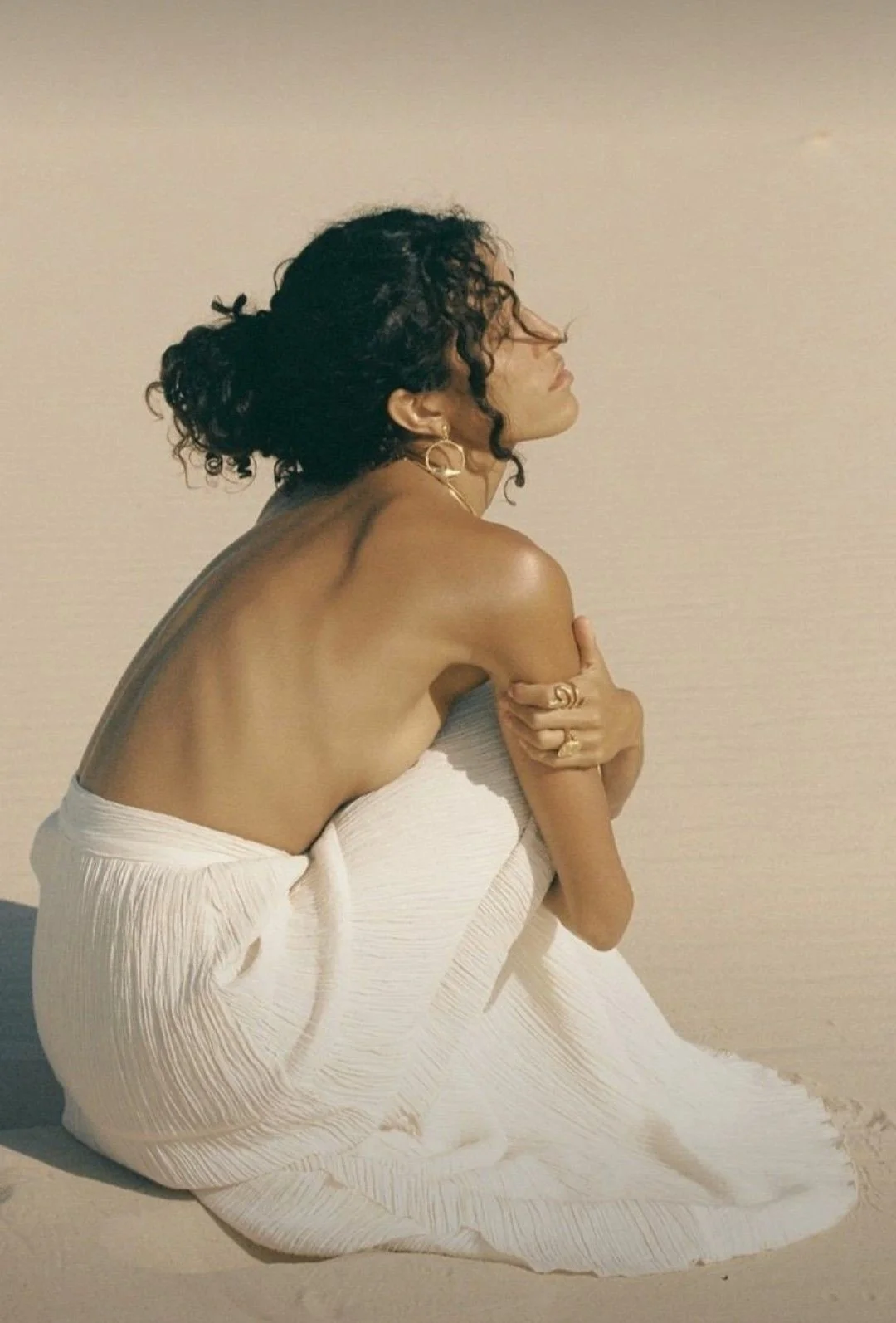 A woman with curly hair sitting on the floor, wrapped in a white fabric, with her arms crossed over her chest and holding her shoulders, against a plain beige wall.